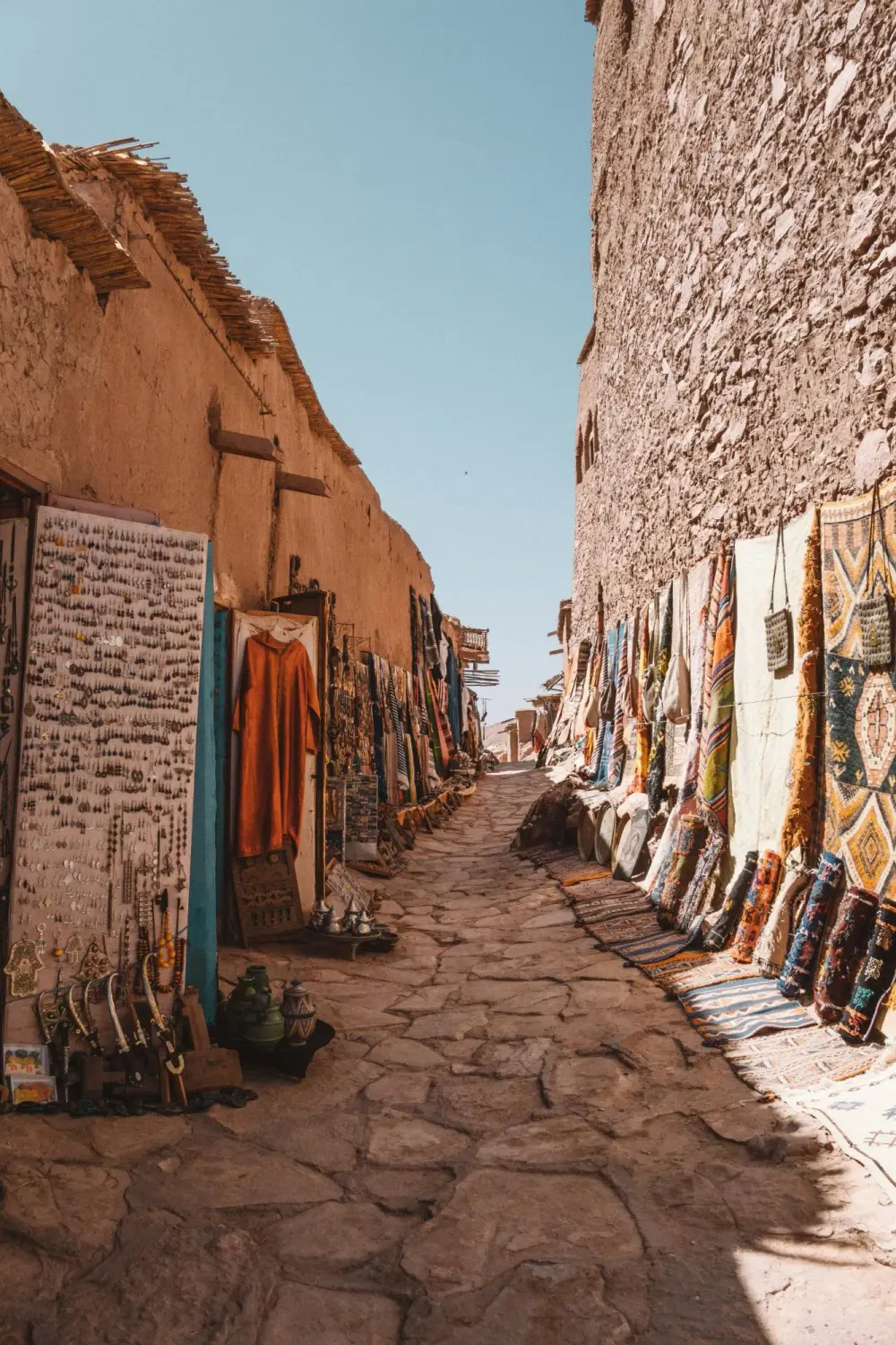 Ruelle pavée bordée de tapis, de bijoux et de tissus dans un ksar du sud du Maroc