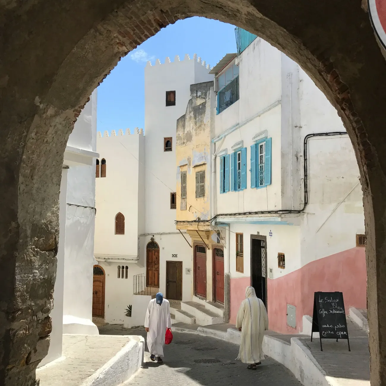 Vicolo bianco della medina di Tangeri visto attraverso un arco, con persiane azzurre e case merlate