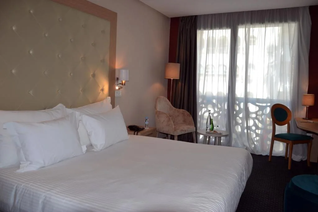 Zimmer mit einem großen weißen Bett, einem gepolsterten Kopfteil und einer bodentiefen Glastür zu einem Balkon