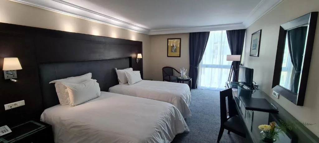 Zimmer mit zwei Einzelbetten, dunklem Kopfteil, Schreibtisch und großen Fenstern