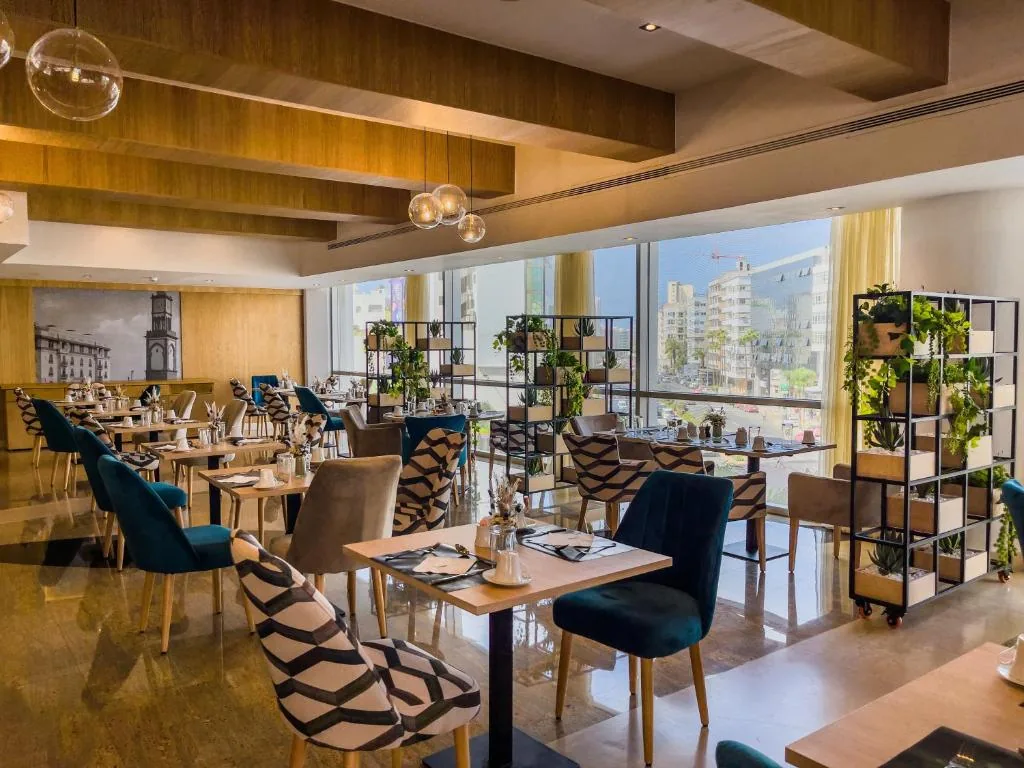 Sala da pranzo luminosa con soffitto a travi di legno, grandi vetrate sulla città, sedie blu petrolio e scaffali di piante