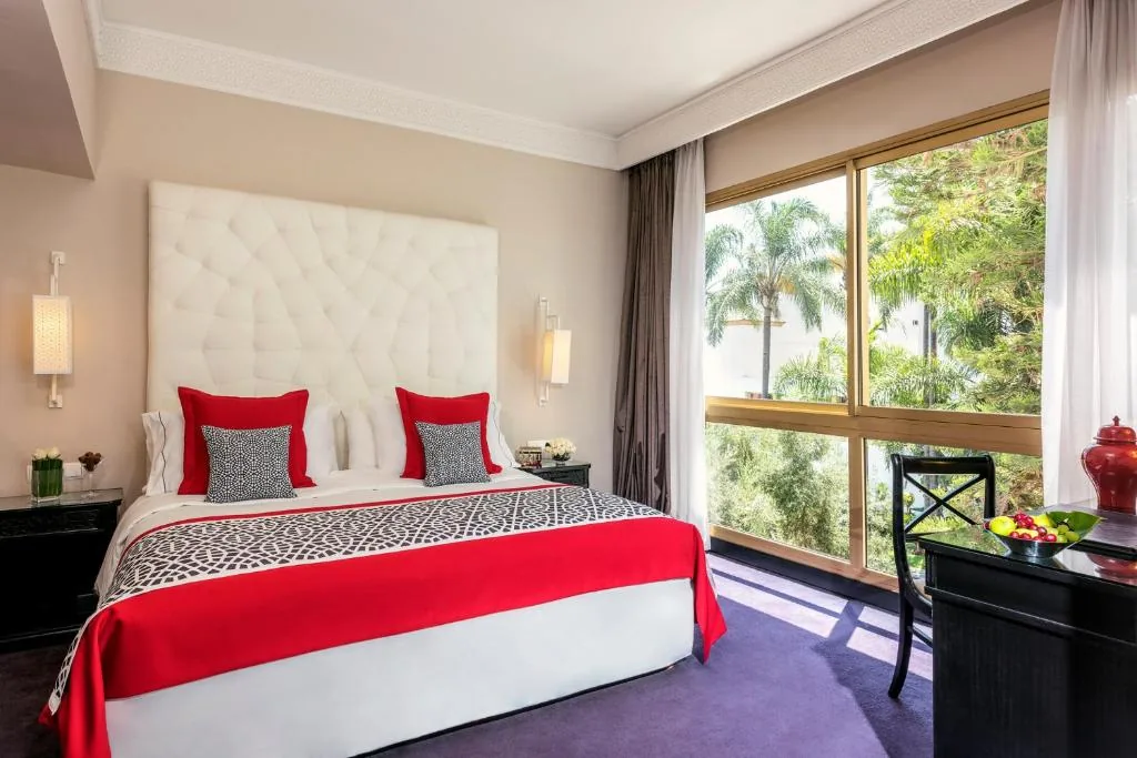 Room with a large bed, a white quilted headboard, a red throw and large picture windows onto palm trees