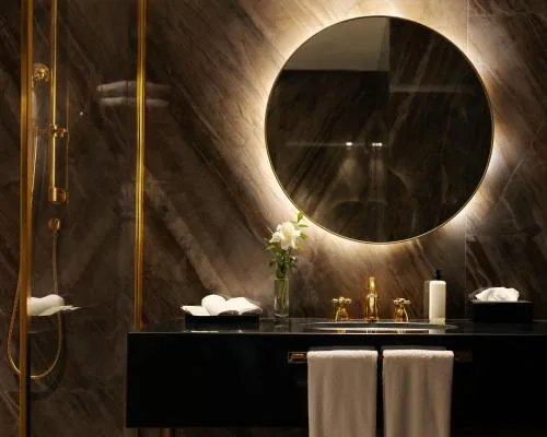 Cuarto de baño con un espejo redondo retroiluminado, una encimera de mármol oscuro y grifería dorada