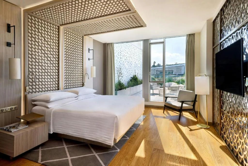 Zimmer mit einem großen weißen Bett, durchbrochenen Holzpaneelen und einem Panoramafenster zu einem Balkon