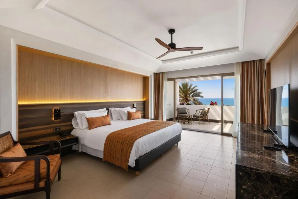 Room with a large bed, a wooden headboard, a ceiling fan and a French window opening onto the sea