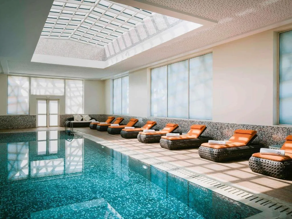 Bassin au carrelage turquoise sous une verrière, bordé de transats en rotin tressé