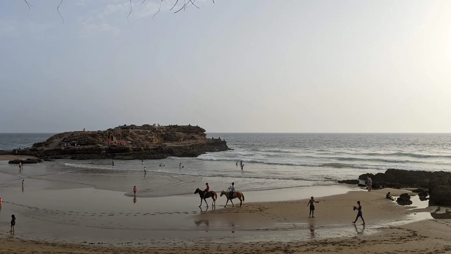 Two riders on horseback along a wide beach, with walkers nearby