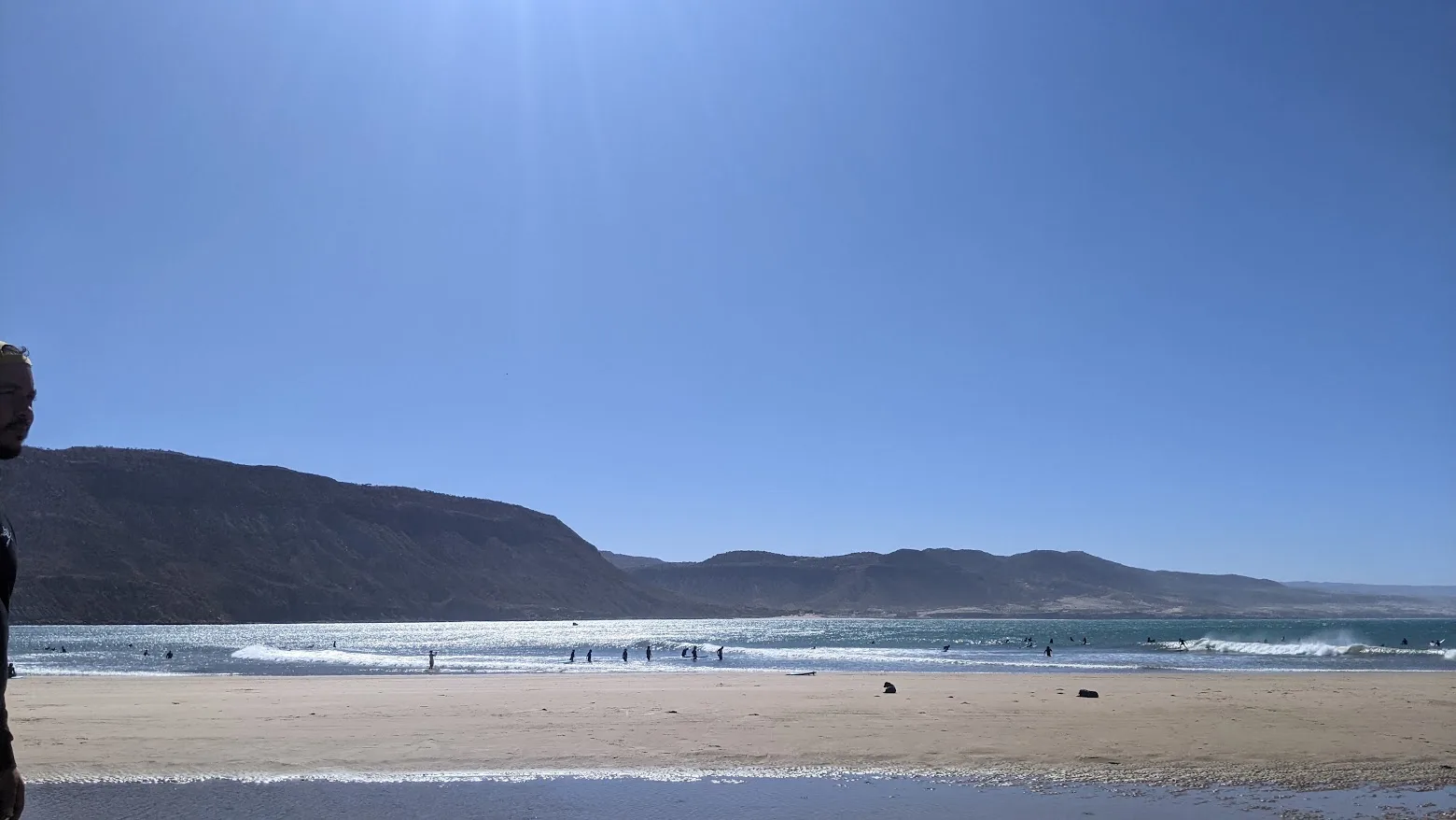 A wide beach beneath a mountain, with waves and a few people in the water