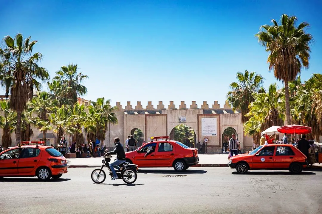 Taxis rouges devant l'entrée crénelée du Souk El Had d'Agadir, entre des palmiers